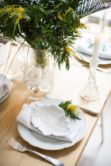Beautiful springtime table setting with green leaves and mimosa branches, bright white table dinner decoration