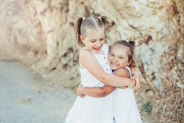 Happy cute laughing girls in white dresses hugging each other on rocks background. Happiness, vacation, support concept