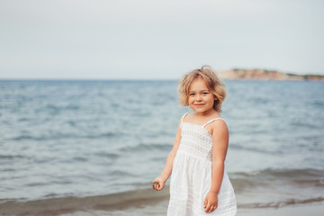 Fototapeta na wymiar Cute adorable little girl in white dress walking on the beach. Happiness, childhood, sea, new generation concept