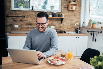 Happy handsome man using laptop in the morning at the kitchen table.