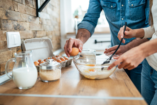 Couple Cooking Together. Adding Eggs Into Glass Bowl With Flour And Mixing In The Kitchen Of The Home.