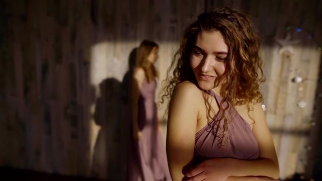 In the foreground is a girl with curly hair and a delicate purple dress. In the background there is a second girl in a similar dress and comes to the first from the back.