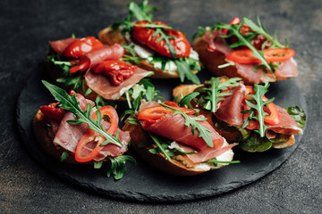 Italian bruschetta with prosciutto, dried tomatoes and arugula on a dark background.