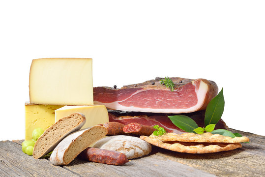 Diverse S&uuml;dtiroler Spezialit&auml;ten isoliert auf einem Holztisch vor wei&szlig;em Hintergrund &ndash; South Tyrolean specialties like bacon, sausages and cheese lying on a rustic table in front of a white background
