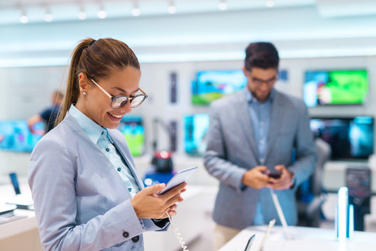 Happy beautiful Caucasian woman with ponytail and dressed in suit buying new smart phone. Tech store interior.