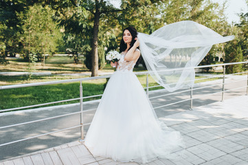 Cute bride in a white dress with a bouquet poses in the garden with greens and thujas. Portrait of a beautiful and smiling brunette with a long veil. Wedding photography.