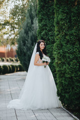 Obraz na płótnie Canvas Cute bride in a white dress with a bouquet poses in the garden with greens and thujas. Portrait of a beautiful and smiling brunette. Wedding photography.