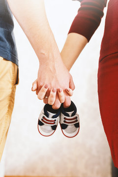 Close Up Of Happy Parents Awaiting Baby And Holding Hands. In Hands Baby Shoes.