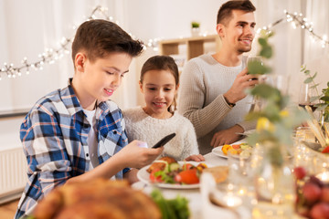 technology, holidays and people concept - happy children with smartphone at family dinner party