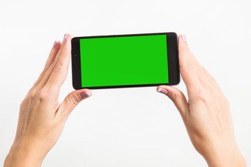 Close up mockup image of woman's two hands holding black mobile phone with blank green screen isolated on white background. Horizontal color photography.