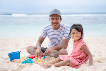 togetherness of father and little girl when look at camera sit on white sand beach