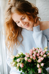 Portrait of a young beautiful girl with a bouquet of roses near window. Curly flowing red hair.