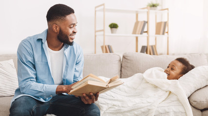 African-american father reading book to daughter on sofa