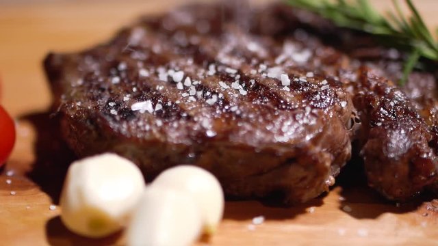 Salt is sprinkled on toasted tasty steak lying on a kitchen board with juicy red tomatoes and onions. Close-up.