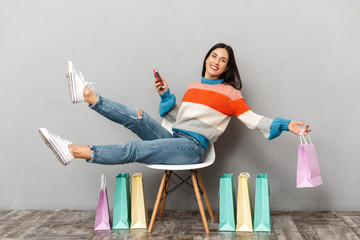 Image of attractive woman 30s sitting in chair with colorful shopping bags and using mobile phone, isolated over gray background