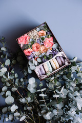Delicious colourful french macaroons cookies in a gift box along with blossoming flower bouquet of roses, mattiola, carnations, eucalyptus on the blue wooden table background with eucalyptus as decor
