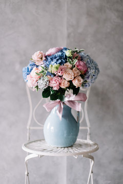 Very nice young woman holding beautiful blossoming bouquet of fresh roses, eustoma, hydrangea, peony, flowers in pink and blue colors in the vase on the grey wall background 