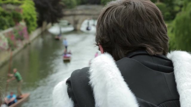 Cambridge University graduate in gown and hood watches punts on River Cam, Cambridge, England