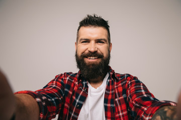 Fototapeta premium cheerful bearded man in checkered shirt taking selfie, isolated on grey