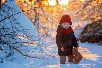 Baby playing with teddy in the snow, winter time. Little toddler boy in blue coat, holding teddy bear on sunset, playing outdoors in winter park