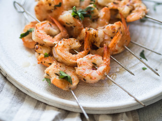 Grilled spicy prawns on metal skewers with parsley and lime juice on white plate on grey background.