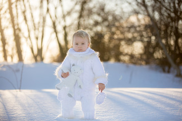 Baby playing with teddy in the snow, winter time. Little toddler boy in handmade white snowsuit, holding teddy bear on sunset, playing outdoors in winter park