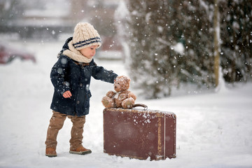 Baby playing with teddy in the snow, winter time. Little toddler boy in blue coat, holding suitcase...