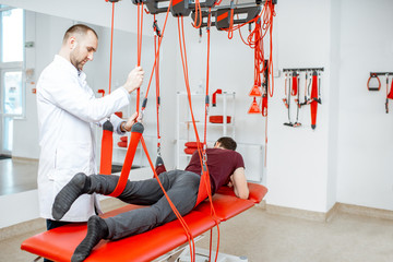 Senior physiotherapist doing treatment to a man with spine problems with suspension medical...