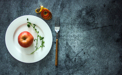 An apple on a white plate next to measuring tape and cutlery, a background about diets, a background about losing weight, the theme of vegetarianism