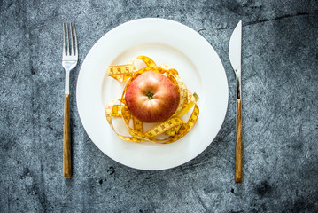 An apple on a white plate next to measuring tape and cutlery, a background about diets, a background about losing weight, the theme of vegetarianism