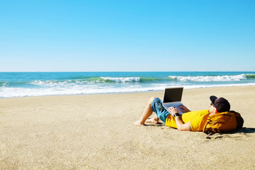 Fototapeta na wymiar Traveler writing blog entry on white laptop, lying at exotic empty beach. Freelance remote work concept. Self employed fit young male in bright yellow t-shirt coding. Copy space, sea view background