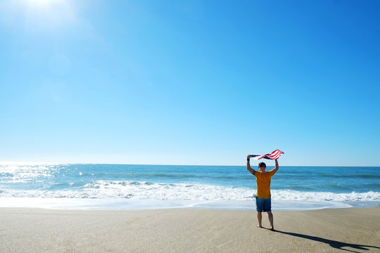 Young athlete running towards ocean holding american flag. Fit surfer man waving United States of America flag. 4th of July Independence day celebration concept. Back view, copy space. - Powered by Adobe