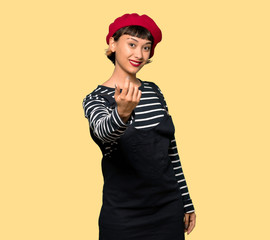 Young woman with beret inviting to come with hand. Happy that you came over yellow background