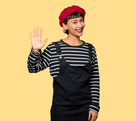 Young woman with beret saluting with hand with happy expression over yellow background