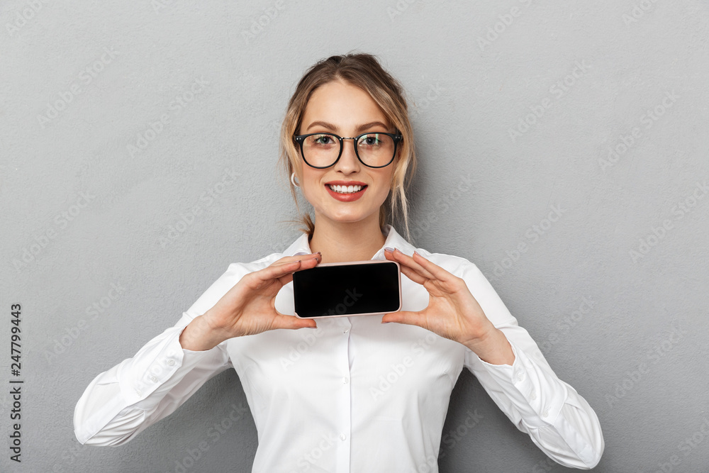 Canvas Prints Portrait of beautiful businesswoman wearing glasses smiling and holding smartphone in the office, isolated over gray background