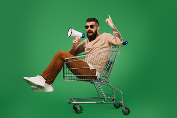 bearded man with megaphone pointing up while sitting in shopping cart isolated on green