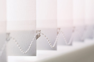 A row of white vertical blinds  cover the sunlight from the window. Close up.