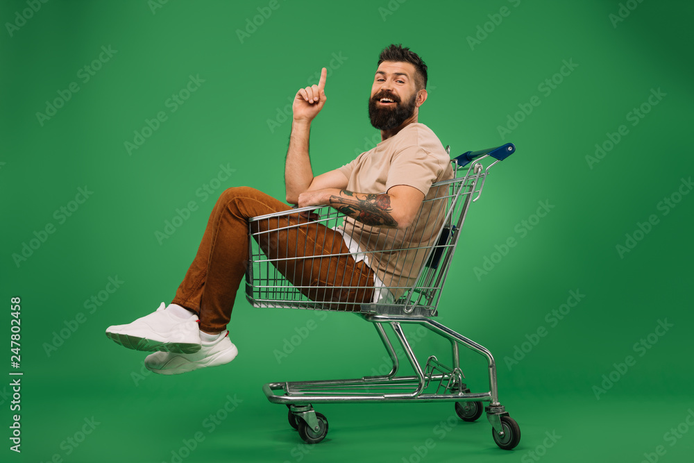 Sticker smiling man pointing up and sitting in shopping cart isolated on green