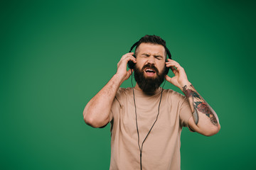 handsome tattooed man singing and listening music in headphones isolated on green