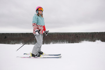 A beautiful young chewing woman in an extreme sports suit, hat and a helmet with a mask on a winter slope is skiing against the background of the forest.