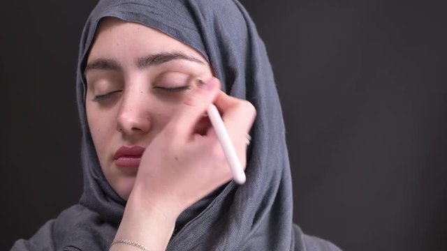 Portrait of female hands doing make-up drawing a line with brown pencil for young muslim woman in hijab on black background.