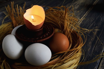 Burning candle in eggshell as symbol of life on ceramic candlestick among eggs in nest of bast on dark old wooden background. Easter concept. Selective focus.