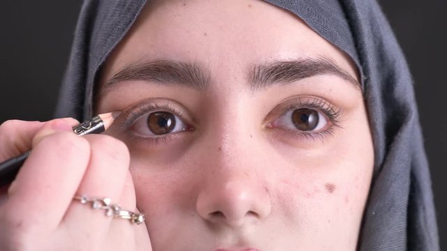 Close-up portrait of female hands doing eye make-up with brown pencil and brush for young muslim woman in hijab on black background.