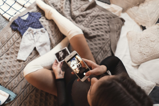 Pregnant young woman taking pictures of ultrasound photos with smartphone while relaxing on a bed