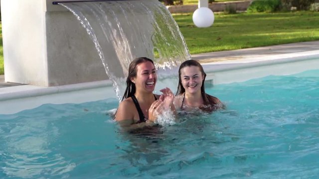 Two beautiful women playing with villa pools waterfall on sunny day.
