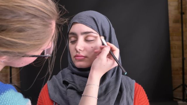 Portrait of female hands doing eye make-up with brown pencil and brush for beautiful muslim woman in hijab on black background.