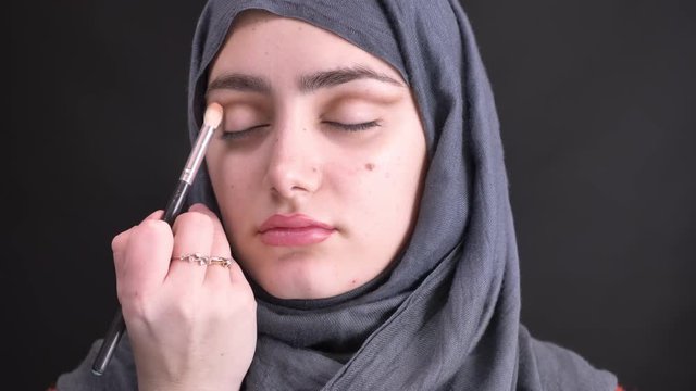 Portrait of female hands doing eye make-up stumping with brush for beautiful muslim woman in hijab on black background.