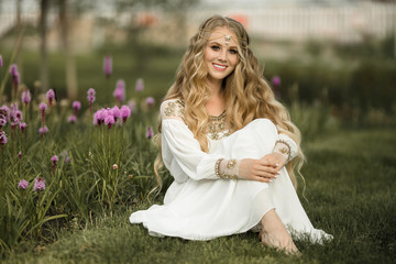 Happy smiling blonde girl with long hair is sitting on grass in spring park with purple flowers