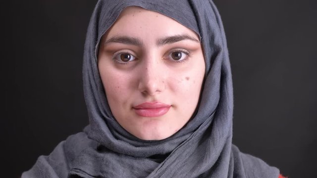 Portrait of young muslim woman in hijab with half-finished make-up watching calmly in camera on black background.