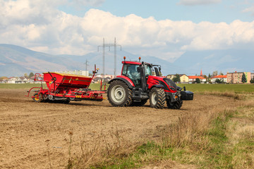 Fototapeta premium Red tractor sowing, pulling seeder trailer behind, on dry field, some houses and mountains in background.